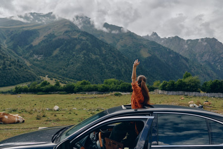 Woman in an open car window raises an arm as mountains tower over a wide landscape, symbolizing travel, adventure, nature and freedom.の写真素材