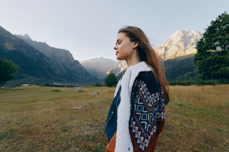 Woman in profile stands in mountain meadow wearing patterned jacket, nature landscape and golden sunlight. Young portrait outdoors, serene hiking scene and peaceful alpine scenery.の写真素材