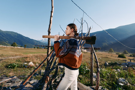 Woman carrying wood on rustic fence in mountains, wearing a warm sweater in a rural landscape; portrait of outdoor activity and traditional lifestyle on a sunny day.の写真素材