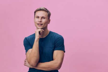 Confident young man with blonde hair, wearing casual blue t-shirt, posing thoughtfully with hand on chin against pink background, studio shot, portrait, people lifestyle concept, modern fashion.の写真素材