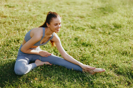Flexible young woman in gray yoga outfit stretching on green grass during a sunny day, showcasing determination and focus in a natural settingの写真素材
