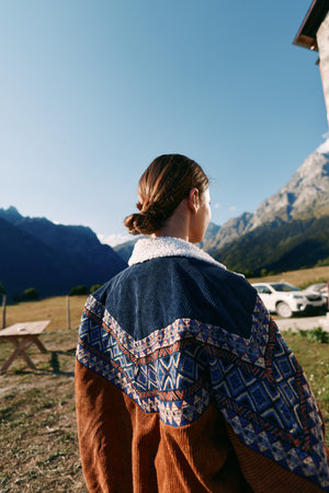 Woman in a patterned sweater stands with her back to camera in the mountains outdoors, overlooking a quiet countryside landscape under clear sky, enjoying autumn light and peaceful nature.の写真素材