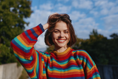Woman smile rainbow sweater portrait outdoors happy young short hair under sunny blue sky, cheerful female in colorful knit, natural light and casual pose for lifestyle portrait.の写真素材