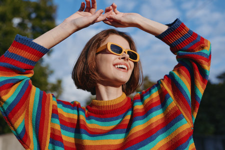 woman in her twenties wearing a colorful striped sweater and yellow sunglasses, smiling and raising hands outdoors in a sunny park, joyful casual outfit lifestyle portraitの写真素材