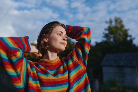 Young woman in a rainbow striped sweater outdoors in a rural setting, smiling with arms raised,...