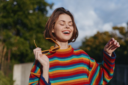 Young woman in her mid 20s smiling woman outdoors, joyful and carefree, wearing a colorful striped sweater and holding yellow sunglasses, casual outfit lifestyle portrait in a sunny park.の写真素材