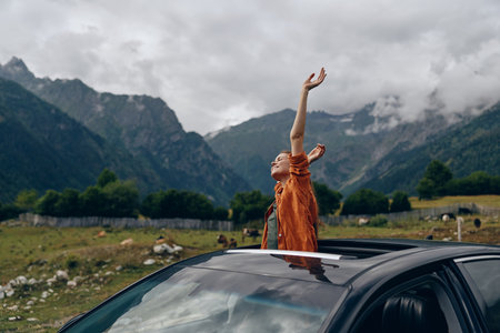 outdoor scene with a person standing through a car sunroof wearing an orange top arms raised amid mountains conveying freedom adventure and joyful travel in natureの写真素材