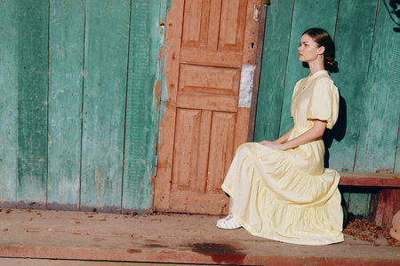 woman sitting on a bench in a flowing yellow dress beside a weathered green wall and wooden door, an outdoor portrait radiating calm, vintage charm, and soft daylightの写真素材