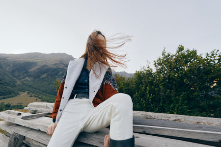Woman sitting on wooden bench in mountains, hair blown by wind across her face, wearing cozy jacket, white pants and boots, relaxed outdoors travel portrait in natural landscape.の写真素材
