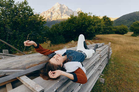 Woman lying relaxed on a wooden fence in a meadow with mountain backdrop and forest, enjoying nature and peaceful outdoor moment in casual outfit during a sunny summer escape.の写真素材