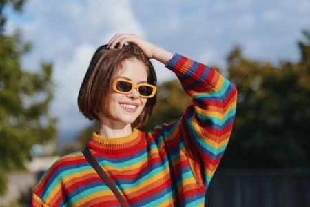 Woman portrait smile in rainbow sweater and yellow sunglasses outdoors, short hair and casual fashion look, cheerful young adult enjoying sunny autumn dayの写真素材