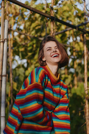 Woman smiling in a colorful rainbow sweater, young adult female laughing outdoors among lush...