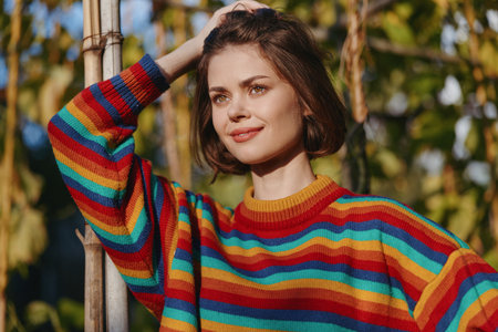 Woman smiling in a colorful striped sweater, short hair portrait leaning on bamboo pole outdoors in a sunlit garden. Casual outfit, relaxed mood for lifestyle and travel.の写真素材