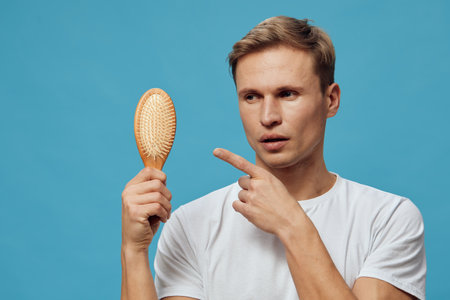 young caucasian man holding hairbrush pointing serious expression blue background casual white...