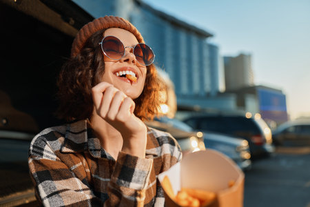 Woman with sunglasses and beanie smiling while eating fries in urban parking at golden hour; candid lifestyle portrait with authenticity, emotional storytelling and mindful living atmosphere.の写真素材