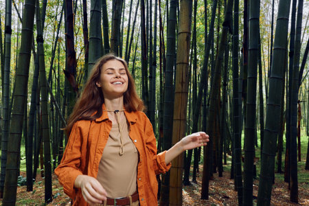 Woman in bamboo forest smiling with eyes closed, happy portrait in nature wearing an orange jacket, peaceful outdoor scene of relaxation and serene sunlight on trunks.の写真素材