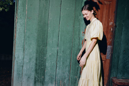 Casual portrait of a woman in a light dress walking beside a weathered doorway in an outdoor setting, with soft daylight and a vintage ambiance.の写真素材