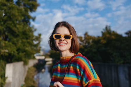 Woman smiling in orange sunglasses and rainbow sweater, portrait outdoors with blue sky, warm sunlight and shoulder bag; happy young adult in colorful casual street style, candid cheerful vibe.の写真素材