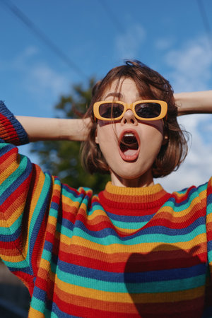 Woman in yellow sunglasses and rainbow sweater surprised with open mouth in a colorful portrait outdoors. Short hair, expressive fashion vibe and playful mood against blue sky.の写真素材