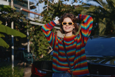 Woman in her mid 20s wearing a colorful striped sweater and yellow sunglasses, smiling and leaning...