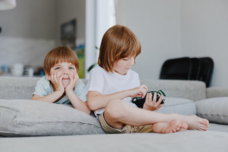Two children playing at home, one boy resting his chin on his hands with a smile, while the other focuses on a toy. A cozy and inviting atmosphere surrounds them.の写真素材