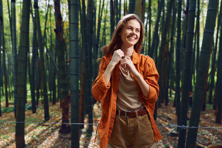 Woman in bamboo forest smiling and enjoying nature outdoors, wearing orange jacket and casual shorts, travel portrait with sunlight filtering through tall bamboo stalks and green canopy.の写真素材