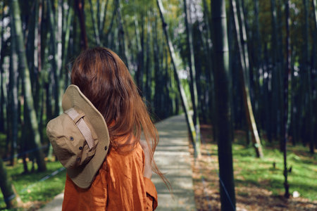 Woman with hat and long hair seen from back walking along a paved path through dense bamboo forest, wearing rust orange coat, travel mood, nature trail, serene peaceful outdoor scene for wanderの写真素材