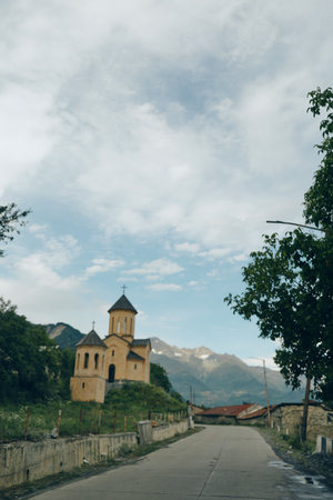 Countryside road leads to a small church set against towering mountains under a bright sky, creating a serene landscape with rural charm and peaceful village atmosphereの写真素材