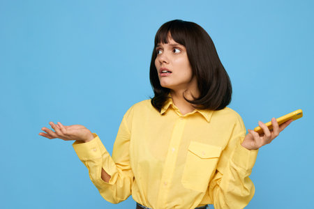 A thoughtful young woman stands against a bright blue backdrop, wearing a yellow shirt, holding a smartphone, and looking puzzled as she questions something on screen.の写真素材