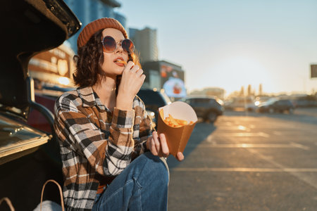Woman eats fries from a car trunk in a parking lot at sunset, wearing sunglasses and a beanie in an urban scene. Authenticity, candid lifestyle and golden hour glow show mindful living.の写真素材