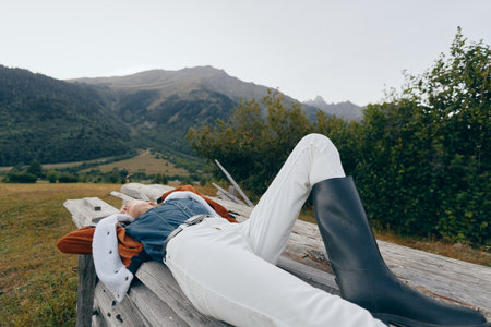 Woman relax on wooden bench in countryside meadow with mountains in background, wearing boots, white pants and denim jacket, outdoor leisure, rural escape and calm nature.の写真素材