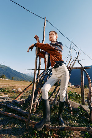 Woman model countryside mountains pose by rustic wooden structure wearing sweater, scarf and boots...