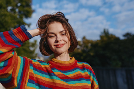 Woman smiling in colorful striped sweater, short hair portrait outdoors in warm sunlight. Young adult model in casual outfit, hand on head, relaxed pose and natural confident lifestyle portrait.の写真素材