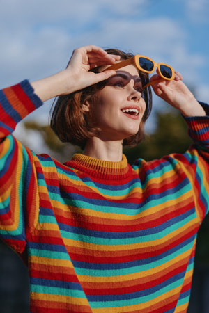 Woman in a colorful striped sweater wearing yellow sunglasses, smile in portrait outdoors while...