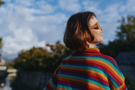Woman in her late twenties wearing a colorful striped sweater and yellow sunglasses smiling and looking up outdoors by riverbank, short brown hair, relaxed travel lifestyle, sunny autumn moodの写真素材