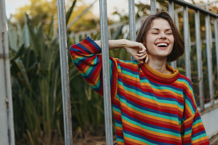 woman smiling in a colorful striped sweater, short hair model leaning on metal fence outdoors, casual outfit, joyful expression, urban garden lifestyle portrait for fashion travelの写真素材