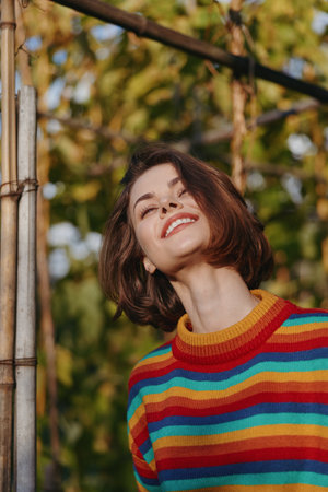 Young woman smiling woman in a colorful striped sweater, short brown hair, outdoor portrait with warm natural light, joyful expression and casual outfit leaning on a wooden pole, lifestyle.の写真素材