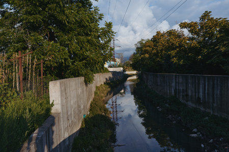 Concrete canal and urban drainage with narrow stream, overgrown vegetation and power lines reflected in water under moody sky at late afternoon, quiet industrial outskirts scene.の写真素材
