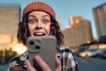 woman phone surprise city beanie selfie. Candid urban portrait of a woman staring at her phone with joyful surprise during golden hour glow, authenticity and emotional storytelling in mindful living.の写真素材