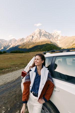 Woman leans on car by mountain road, smiling in warm jacket during outdoor travel and adventure. Scenic landscape with clear sky, relaxed pose, nature exploration and sunlight.の写真素材