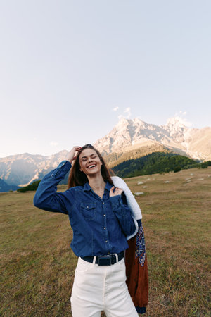Woman with smile in denim shirt and white pants standing in mountains meadow, holding a jacket over...