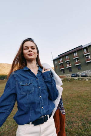Woman smiling in denim shirt holding a jacket outdoors in a grassy field near a residential...