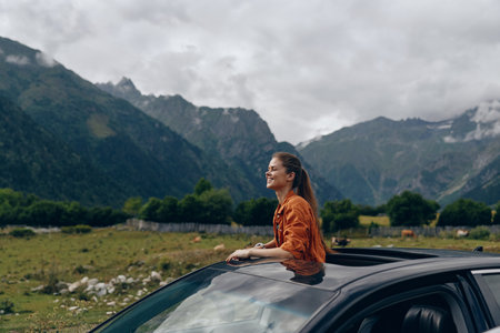 Woman leans from car window, smiling, enjoying an outdoor adventure in a mountainous landscape with open fields and dramatic clouds overhead under blue skiesの写真素材
