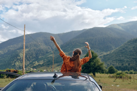 Woman stands on a car roof with arms raised enjoying freedom during a road trip in a scenic mountain landscape outdoors, conveying adventure and happiness on the open roadの写真素材