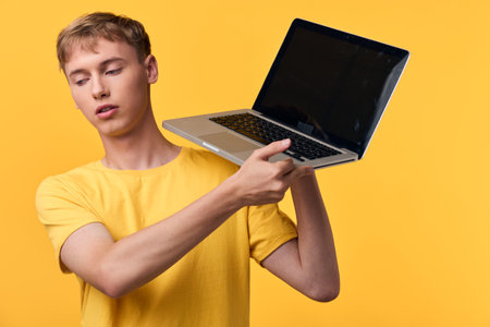 Male in yellow t shirt holding a laptop against a vibrant yellow background, emphasizing portable technology, digital work, and casual online learning in a modern studio lifestyle setupの写真素材