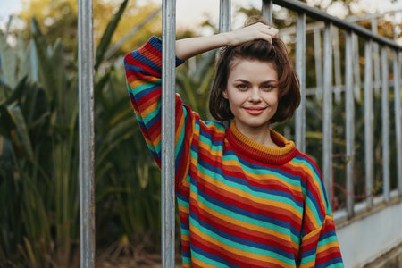 Woman portrait in a colorful sweater smiling and leaning on a metal fence outdoors. Casual short hair, relaxed pose with autumn foliage background, natural light and warm mood.の写真素材