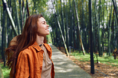 Woman with a smile and redhead hair wearing an orange jacket walking along a bamboo forest path, enjoying nature and sunlight in a peaceful outdoor portrait.の写真素材