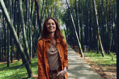 Woman in orange jacket walking along a bamboo path, smiling in a casual portrait style on a paved trail through green forest, nature outdoor scene and relaxed lifestyle.の写真素材