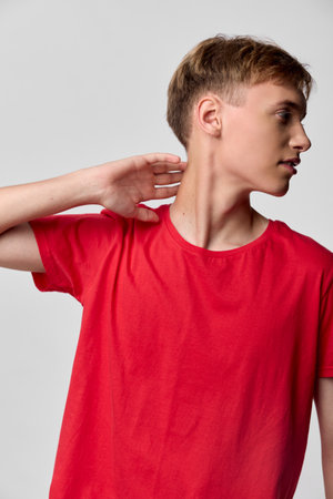 Young man in a red t shirt tilts his head to the side and rests a hand at the neck, posing in a...