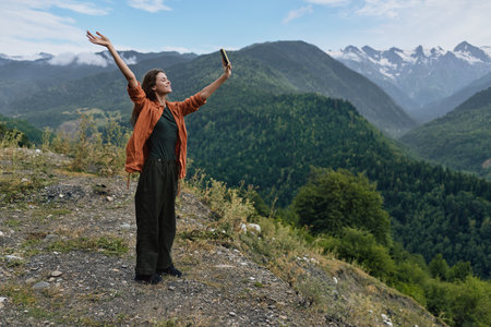 Joyful woman stands on a rocky overlook with arms raised, enjoying panoramic mountains, green...